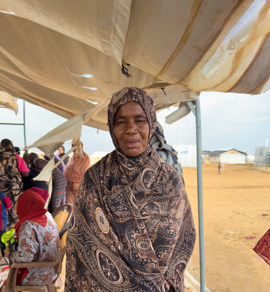 A woman stands under an open-walled tent in an arid village. There are other women in groups behind her.
