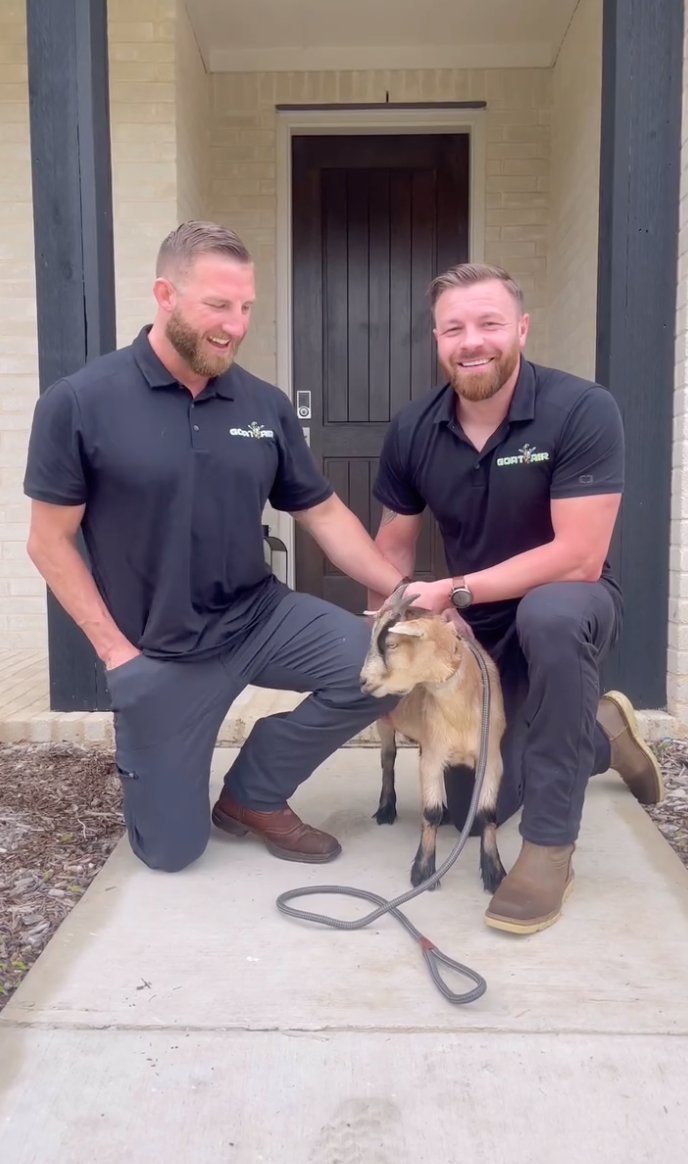 Two men kneel in front of a door. They are petting a goat.