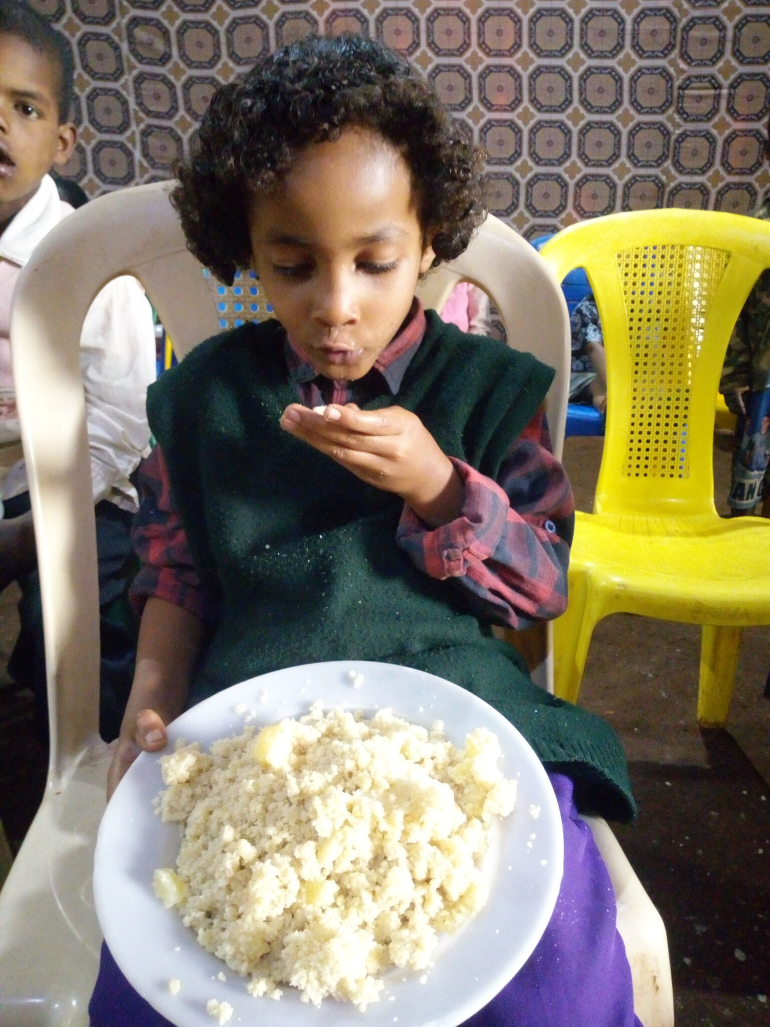 A child is sitting in a chair with a plate full of food. The child is blowing on a handful of the food to cool it down.