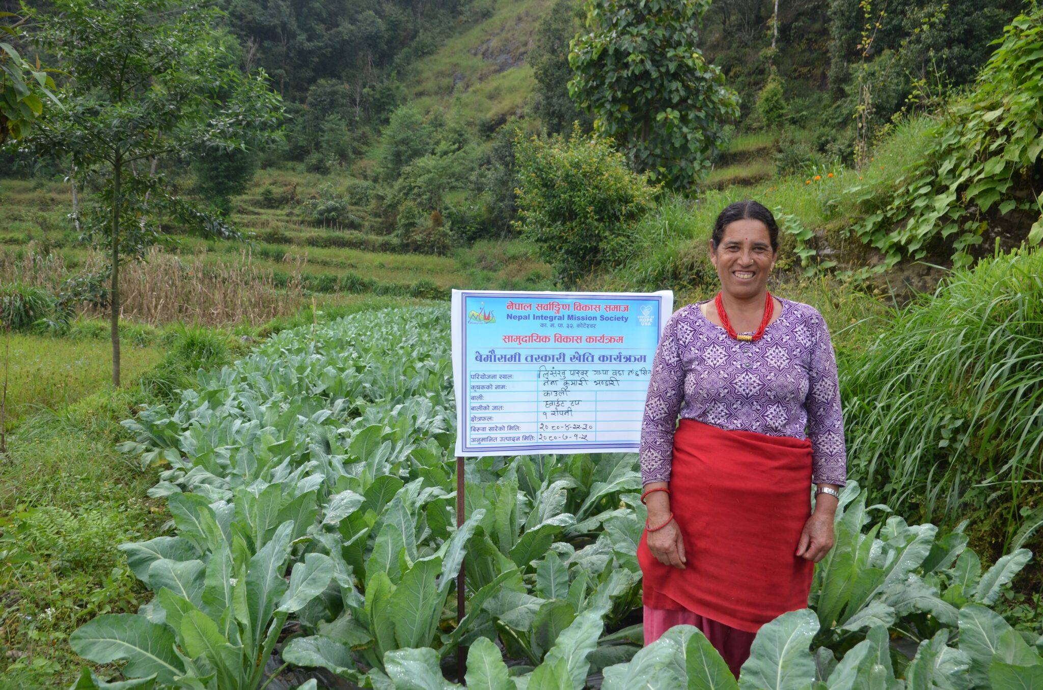 A woman stands proudly among a crop of greens that she has grown.