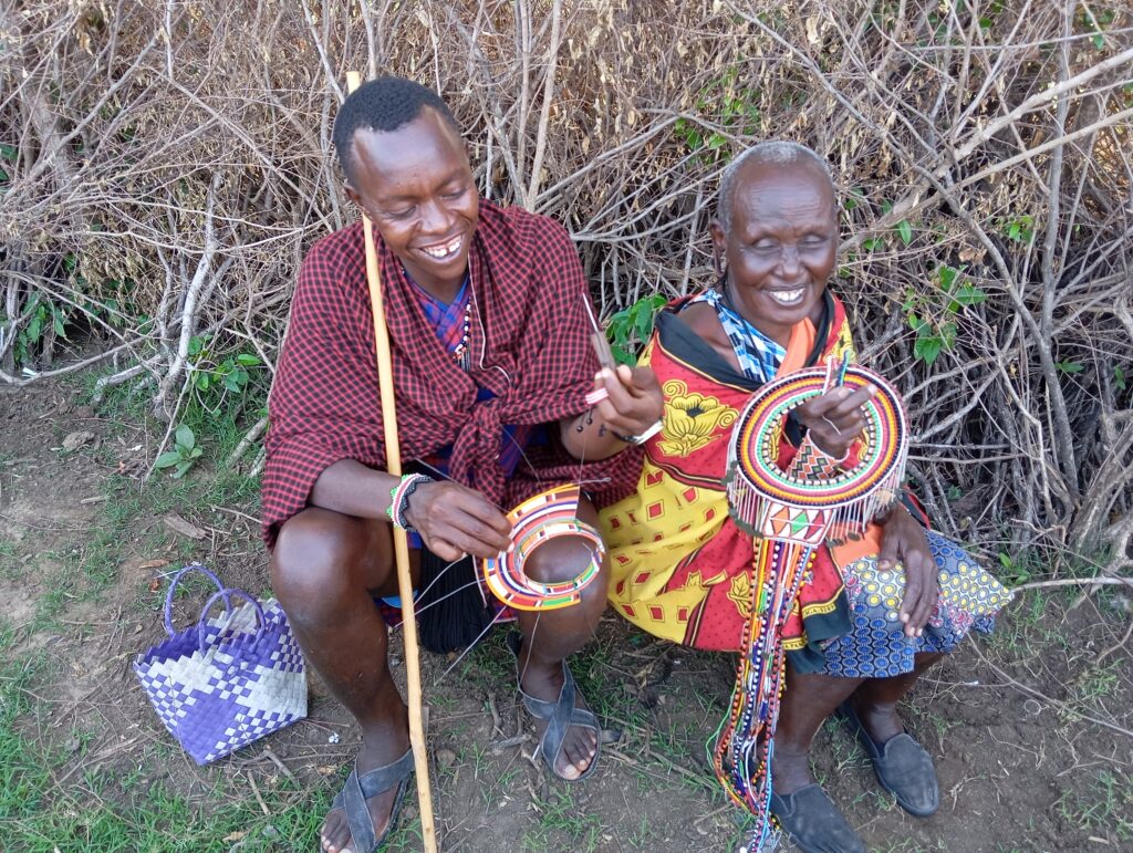 A mother and her eldest son sit next to each other on the ground, making beaded and woven jewelry.